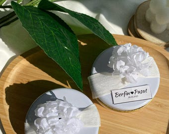 Puede incluir: Dos cajas redondas blancas de regalo con ribete dorado, cada una decorada con una flor blanca y una cinta blanca. Las cajas tienen una etiqueta blanca con texto negro que dice "Berfin ♥ Pusat 18.01.2025".