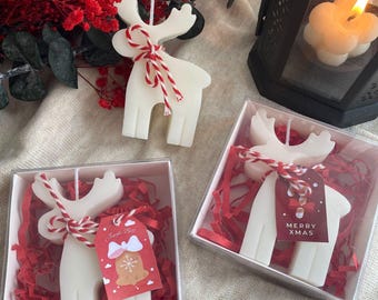 May include: White reindeer-shaped candles with red and white twine bows. Two candles are in clear boxes with red shredded paper and gift tags that say "Merry Xmas" and "Jingle Bell". A lit flower-shaped candle and red flowers are in the background.