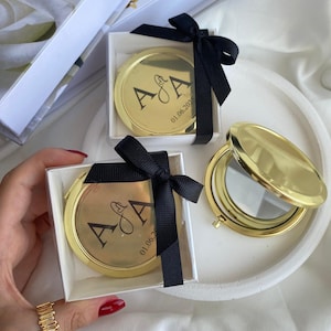 Peut inclure: Miroirs compacts dorés avec initiales et date personnalisées, présentés dans des boîtes blanches avec un ruban noir. Un miroir est ouvert, révélant la surface réfléchissante. Les initiales "A & A" et la date "01.06.202" sont imprimées.
