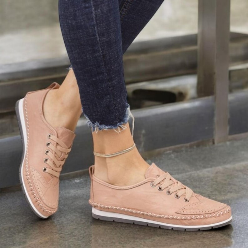May include: A pair of blush-colored sneakers with a lace-up design. The shoes have a white sole with a gray bottom and decorative stitching around the edges. The sneakers are worn with dark blue jeans.
