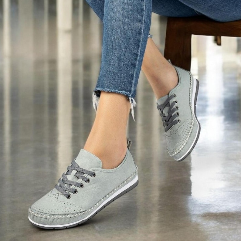 May include: Gray leather sneakers with gray laces and a white sole. The shoes have a classic design with decorative perforations on the toe. The sneakers are shown with blue jeans.