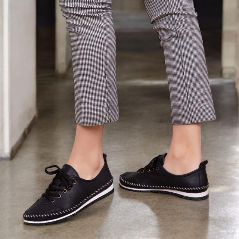 May include: Black leather sneakers with white soles and white stitching. The shoes have laces and are worn with checkered pants. The sneakers are a casual style.