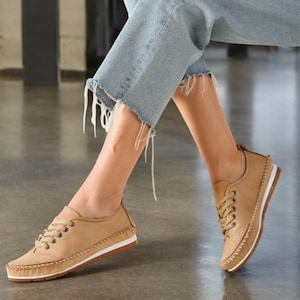 May include: Tan leather sneakers with white soles and brown stitching. The shoes have a lace-up design and a perforated detail on the toe. The sneakers are worn with light blue denim jeans.