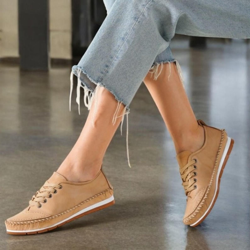 May include: Tan leather sneakers with white soles and brown stitching. The shoes have a lace-up design and a perforated detail on the toe. The sneakers are worn with light blue denim jeans.