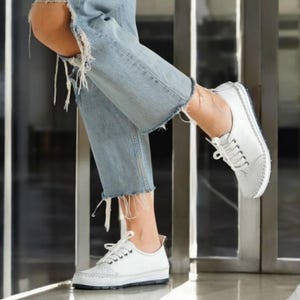 May include: White leather lace-up shoes with a low profile and a stitched sole. The shoes have a classic design with decorative perforations on the toe. The image shows the shoes paired with light blue denim jeans with distressed details.