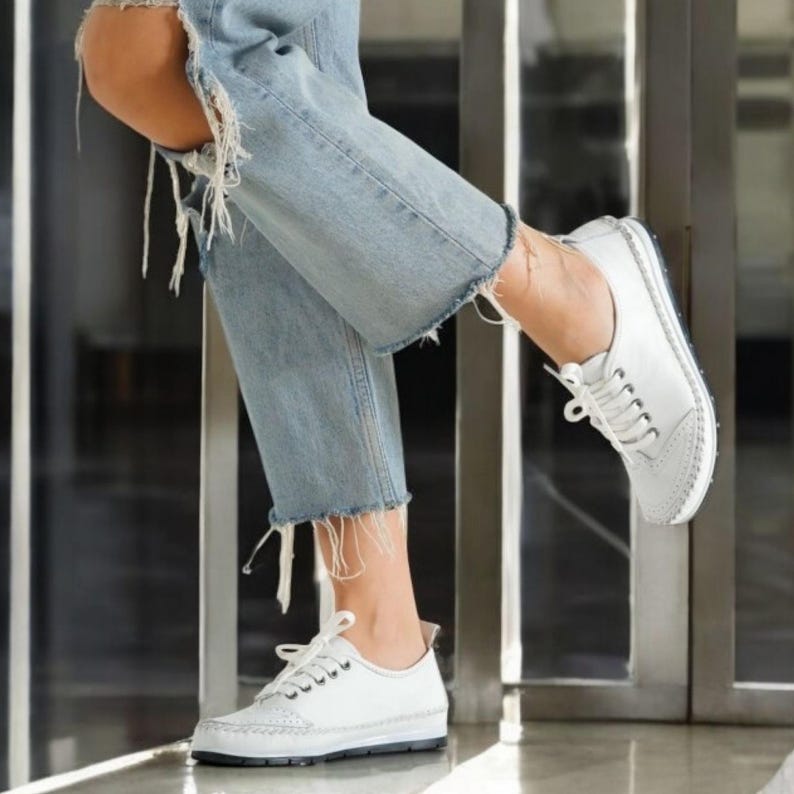 May include: White leather lace-up shoes with a low profile and a stitched sole. The shoes have a classic design with decorative perforations on the toe. The image shows the shoes paired with light blue denim jeans with distressed details.