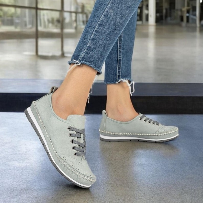 May include: Light gray lace-up sneakers with white soles and gray laces. The shoes have decorative stitching and a perforated design on the toe. The sneakers are paired with blue denim jeans.