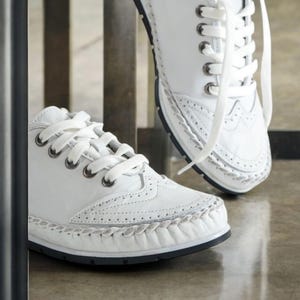 May include: White leather sneakers with decorative stitching and lace-up closure. The shoes have a classic design with a perforated pattern on the toe and sides. The soles are black.