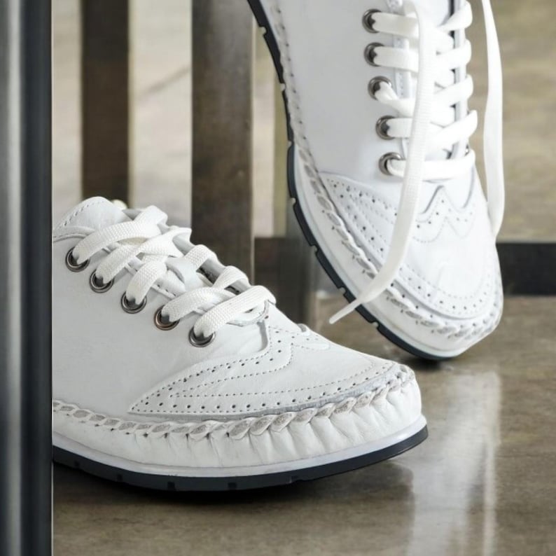 May include: White leather sneakers with decorative stitching and lace-up closure. The shoes have a classic design with a perforated pattern on the toe and sides. The soles are black.