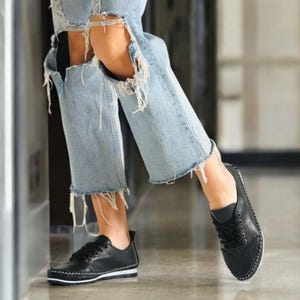 May include: Black leather sneakers with white stitching and soles. The shoes have black laces and a low-profile design. The sneakers are worn with light blue ripped jeans.