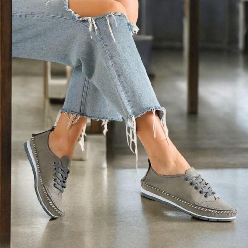 May include: Gray lace-up sneakers with white soles and decorative stitching. The shoes have a low profile and a classic design. The sneakers are shown with light blue ripped jeans.