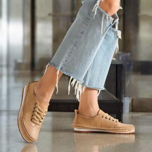 May include: Tan leather sneakers with white soles and decorative stitching. The shoes have a classic lace-up design and a subtle wingtip-style detail on the toe. The sneakers are worn with light blue denim jeans with distressed details.