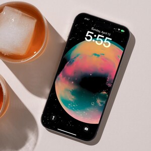 May include: A smartphone with a black background and a colourful planet-like image. The time is 5:55 and the date is Sunday, April 13. The phone is on a white surface.