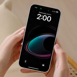 May include: A white smartphone with a black screen displaying the time 2:00 and the date Wednesday, April 2. The background is a dark space scene with a blue and green planet and pink and white rings.