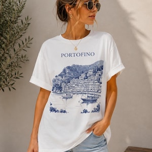 Può includere: T-shirt bianco con un'illustrazione blu di Portofino, Italia, con edifici, barche e acqua. La parola "Portofino" è stampata sopra l'immagine. La maglietta ha maniche corte e una vestibilità rilassata.