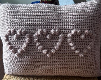 May include: A soft, pink crocheted pillow with three heart-shaped designs created with raised stitches.