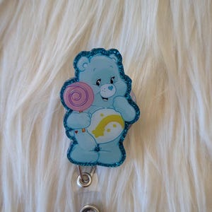 May include: A light blue bear badge reel featuring a glittery border and a graphic of a cartoon bear holding a pink lollipop. The bear has a white belly patch with a yellow crescent moon and star design. A clear plastic strap and metal snap are attached.