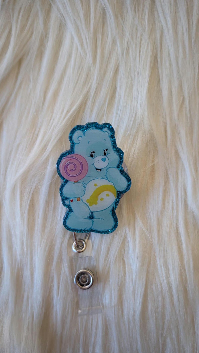 May include: A light blue bear badge reel featuring a glittery border and a graphic of a cartoon bear holding a pink lollipop. The bear has a white belly patch with a yellow crescent moon and star design. A clear plastic strap and metal snap are attached.