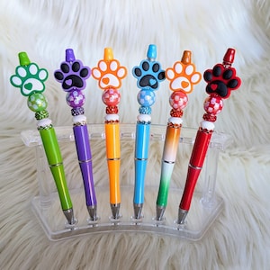May include: A set of six colorful pens with paw print toppers. Each pen has a different color barrel, including green, purple, orange, blue, and red. The paw print toppers are black, white, and various shades of orange and red. The pens are displayed in a clear plastic holder.