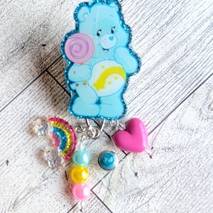 May include: A light blue Care Bear badge reel with a glittery outline, holding a lollipop. Additional charms include a rainbow, a pink heart, and colorful beads. The badge reel is designed for attaching to clothing or accessories.