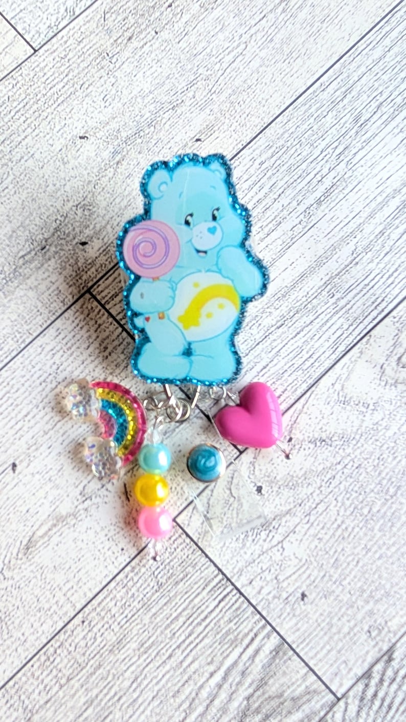 May include: A light blue Care Bear badge reel with a glittery outline, holding a lollipop. Additional charms include a rainbow, a pink heart, and colorful beads. The badge reel is designed for attaching to clothing or accessories.