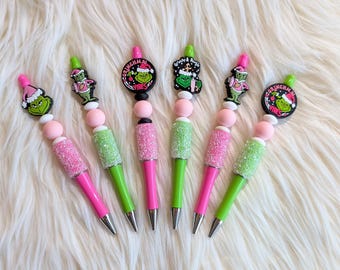 Può includere: Sei penne colorate con decorazioni a tema Grinch. Le penne hanno fusti glitterati rosa e verdi, dettagli rosa e verdi e punte argentate. Le decorazioni presentano il personaggio del Grinch e disegni a tema festivo. Le penne sono disposte su una superficie bianca soffice.