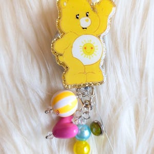 May include: A yellow Care Bear badge reel with a glittery outline and a smiling sun on its belly. The badge reel has a charm of colorful beads in pink, yellow, blue, and green. The background is a fluffy white surface.