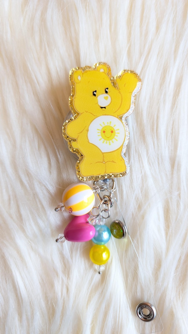 May include: A yellow Care Bear badge reel with a glittery outline and a smiling sun on its belly. The badge reel has a charm of colorful beads in pink, yellow, blue, and green. The background is a fluffy white surface.