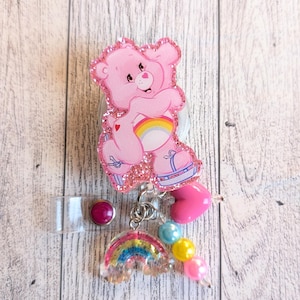 May include: A pink Care Bear badge reel with glitter accents. The bear is holding a rainbow. The reel has a clear plastic clip, a pink heart charm, and a rainbow charm with blue, yellow, and pink beads.
