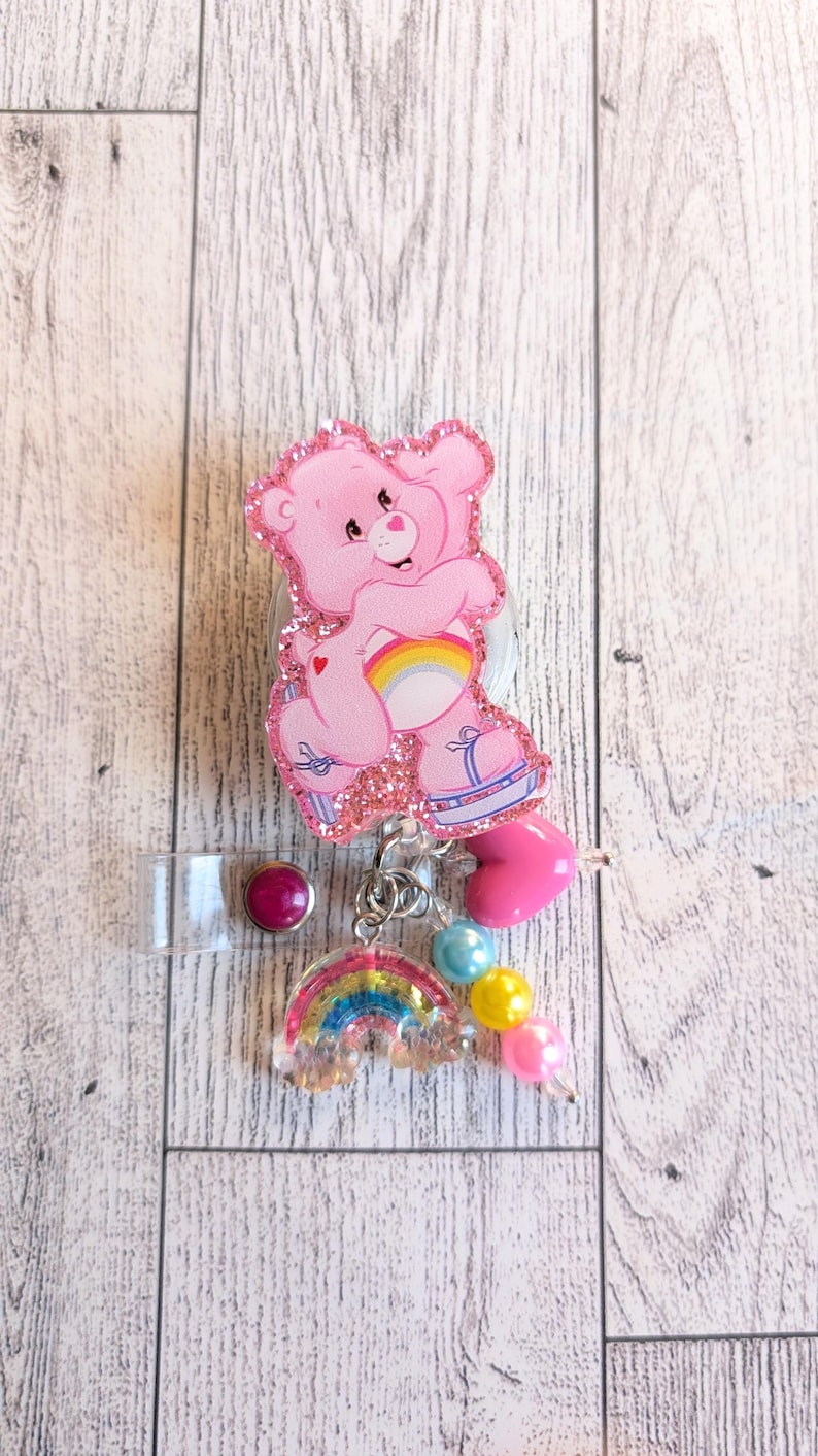 May include: A pink Care Bear badge reel with glitter accents. The bear is holding a rainbow. The reel has a clear plastic clip, a pink heart charm, and a rainbow charm with blue, yellow, and pink beads.