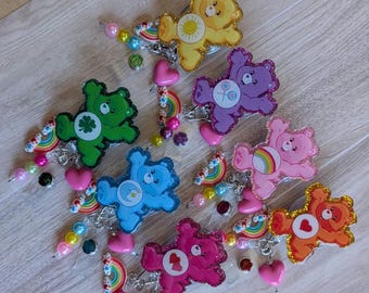 May include: A collection of seven glittery bear-shaped badge reels in yellow, purple, green, blue, pink, orange, and magenta. Each bear features a unique belly symbol and is decorated with dangling charms including small rainbows, beads, and hearts.