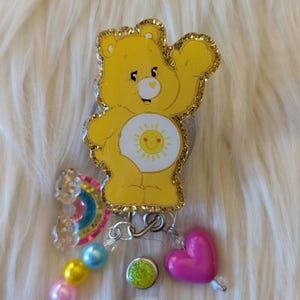 May include: A yellow bear badge reel featuring a sun emblem on its belly, outlined in gold glitter. The reel is decorated with a small rainbow charm, a pink heart bead, and a strand of light blue, yellow, and pink spherical beads.