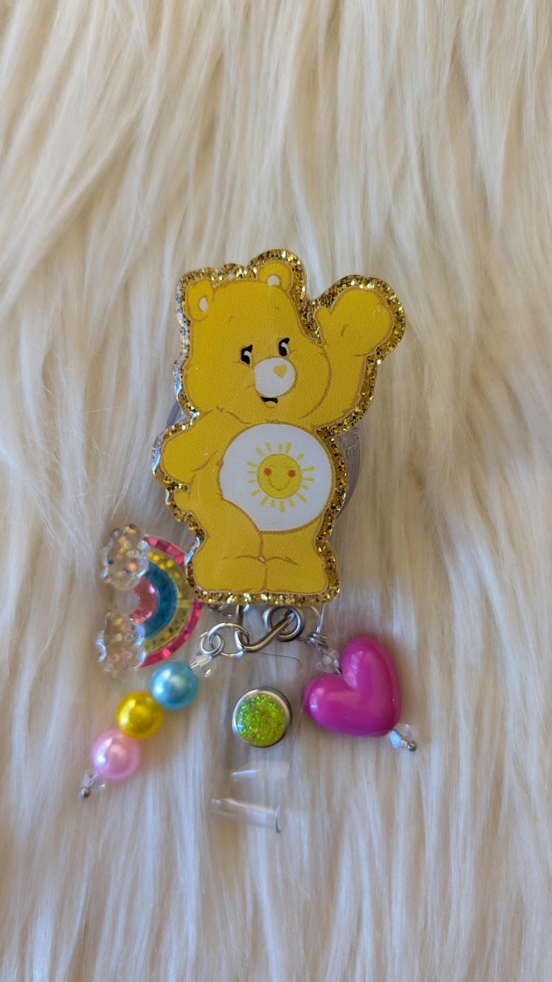 May include: A yellow bear badge reel featuring a sun emblem on its belly, outlined in gold glitter. The reel is decorated with a small rainbow charm, a pink heart bead, and a strand of light blue, yellow, and pink spherical beads.
