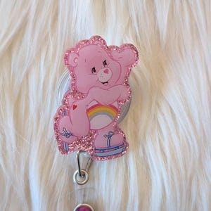 May include: A pink glittery badge reel featuring a cartoon bear character with a rainbow on its belly, wearing blue roller skates. The badge reel has a silver metal clip and a clear plastic strap with a small, round, dark pink decorative stud.