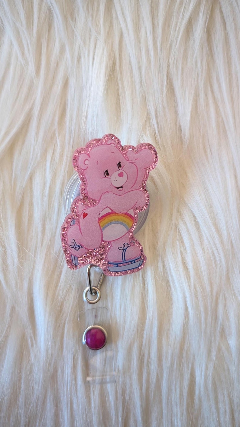May include: A pink glittery badge reel featuring a cartoon bear character with a rainbow on its belly, wearing blue roller skates. The badge reel has a silver metal clip and a clear plastic strap with a small, round, dark pink decorative stud.