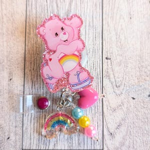 May include: A pink Care Bear badge reel with a glittery outline. The reel features a rainbow, heart, and bead accents. The badge reel has a clear plastic strap and a purple button.