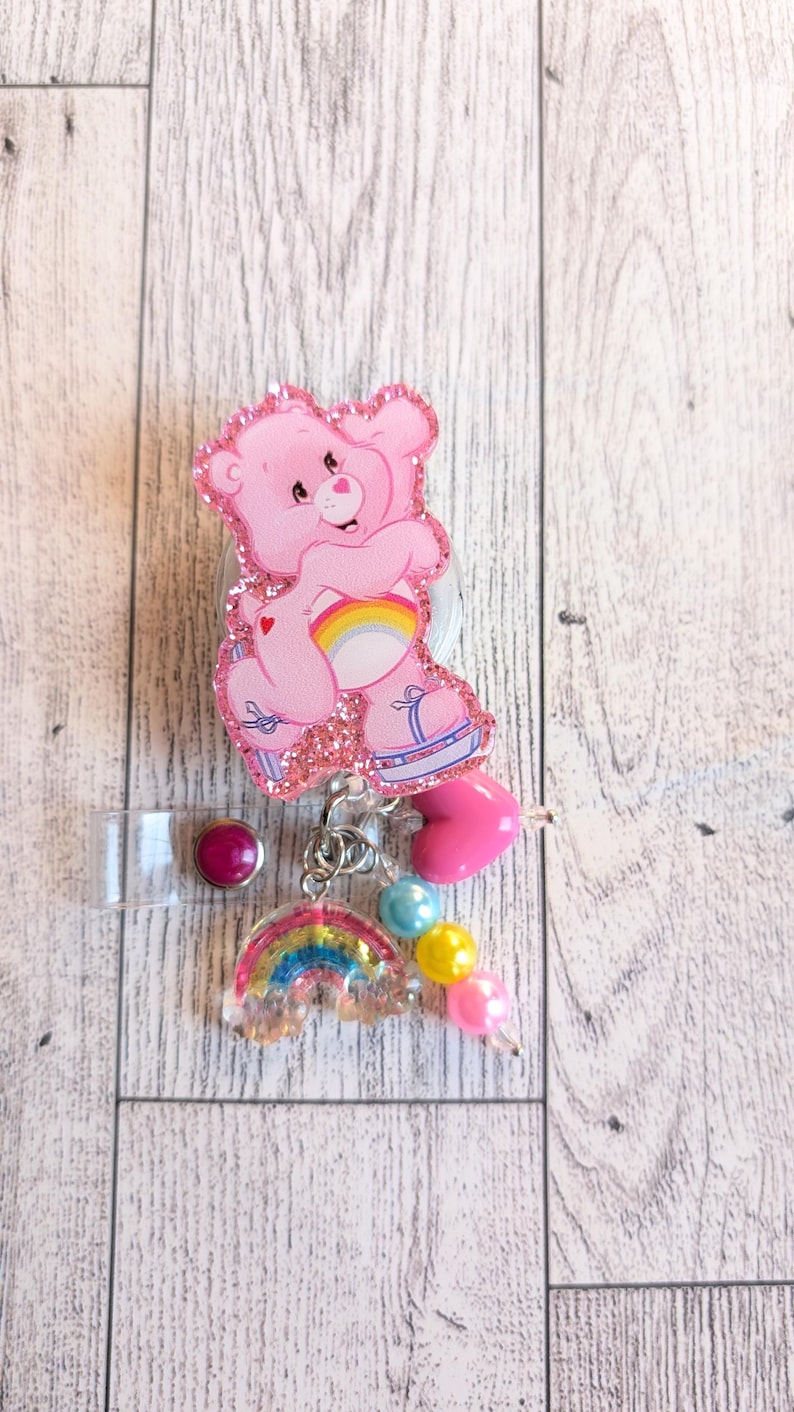 May include: A pink Care Bear badge reel with a glittery outline. The reel features a rainbow, heart, and bead accents. The badge reel has a clear plastic strap and a purple button.