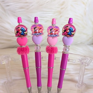 May include: Four decorative pens with pink and purple accents. Each pen features a character design at the top, a heart-shaped bead, and a fluffy pink accent. The pens are displayed on a clear stand against a white, fluffy background.