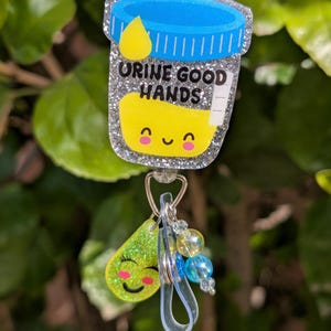 May include: A silver glitter badge with a cartoon urine sample container with a smiling face and the text "Urine Good Hands". The badge has a clear plastic retractable badge holder with a green glitter charm of a smiling raindrop.