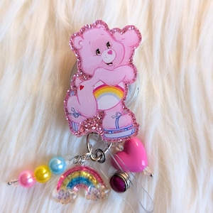 May include: A pink Care Bear-themed retractable badge reel with a glittery finish. The badge reel features a pink Care Bear, a rainbow, a pink heart, and colorful beads. The design is set against a fluffy white background.