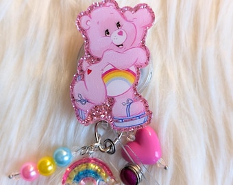 May include: A pink Care Bear-themed retractable badge reel with a glittery finish. The badge reel features a pink Care Bear, a rainbow, a pink heart, and colorful beads. The design is set against a fluffy white background.