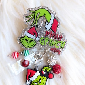 May include: A Christmas-themed badge reel featuring the Grinch. The design includes a glittery background with the words "Merry Grinchmas" and a green Grinch face wearing a Santa hat. Additional decorative beads are attached.