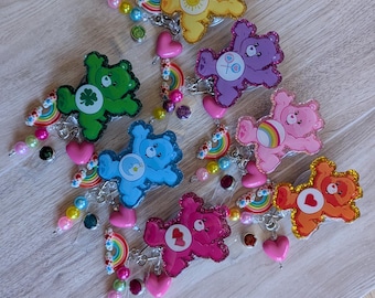 May include: A collection of seven glittery Care Bear badge reels, each featuring a different character in yellow, purple, green, blue, pink, orange, and magenta. Each reel is decorated with colorful beads, small rainbow charms, and heart-shaped accents.
