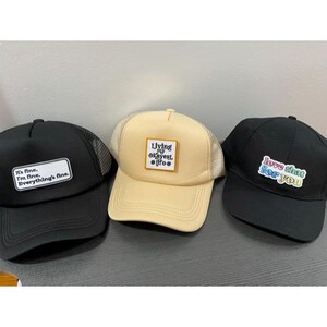 May include: Three trucker hats with different designs. One black hat has a white rectangle with the text "It's fine. I'm fine. Everything's fine." A tan hat has a square patch with the text "Living my okayest life." A black hat has the text "love that for you."