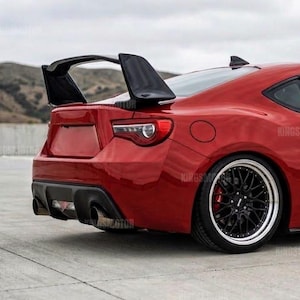 Può includere: Una coupé sportiva rossa a due porte dotata di un grande alettone posteriore nero, cerchi neri a razze multiple con bordi lucidi e un diffusore posteriore. Il veicolo presenta un profilo aerodinamico con vetri oscurati e un assetto ribassato.