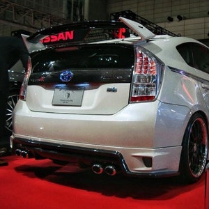 以下が含まれることがあります： 大きなリアウイング、黒いサイドスカート、4本のマフラーエンドを備えたカスタムリアバンパーを備えた白いトヨタプリウス。車にはリアに「DAMD」のロゴがあります。