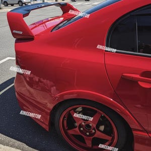 May include: A red car with a black spoiler and red alloy wheels. The car has the text "KINGS MOTOR" in white on the side. The car is parked on a street.