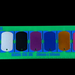 May include: A green plastic display rack holding nine rectangular metal dog tags in various colors including black, purple, white, blue, gold, and red. The rack features the text SONDAWNDESIGNS and DOG TAG 30mm x 50mm printed on its surface.