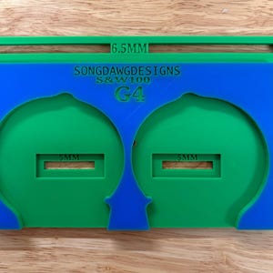 May include: A layered, multi-colored tool with a green frame, a blue center, and green cutouts. The blue layer has the text "SONGDAWD DESIGNS S&W100 G4". Measurements of 16.5mm and 5mm are visible.