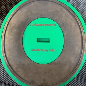 May include: A circular green template with a dark gray textured center insert. The green surface features the red text "SONGDAWGDESIGNS" and "180MM X 203 MM" with a rectangular cutout in the middle. The object has a distinct outer green ring.